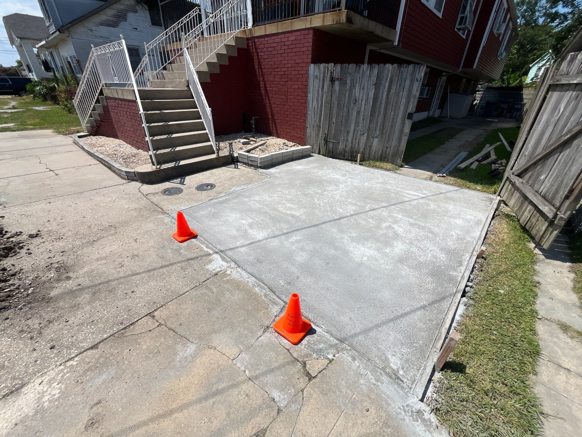 Newly poured concrete pad next to a red brick building with stairs, two orange cones.