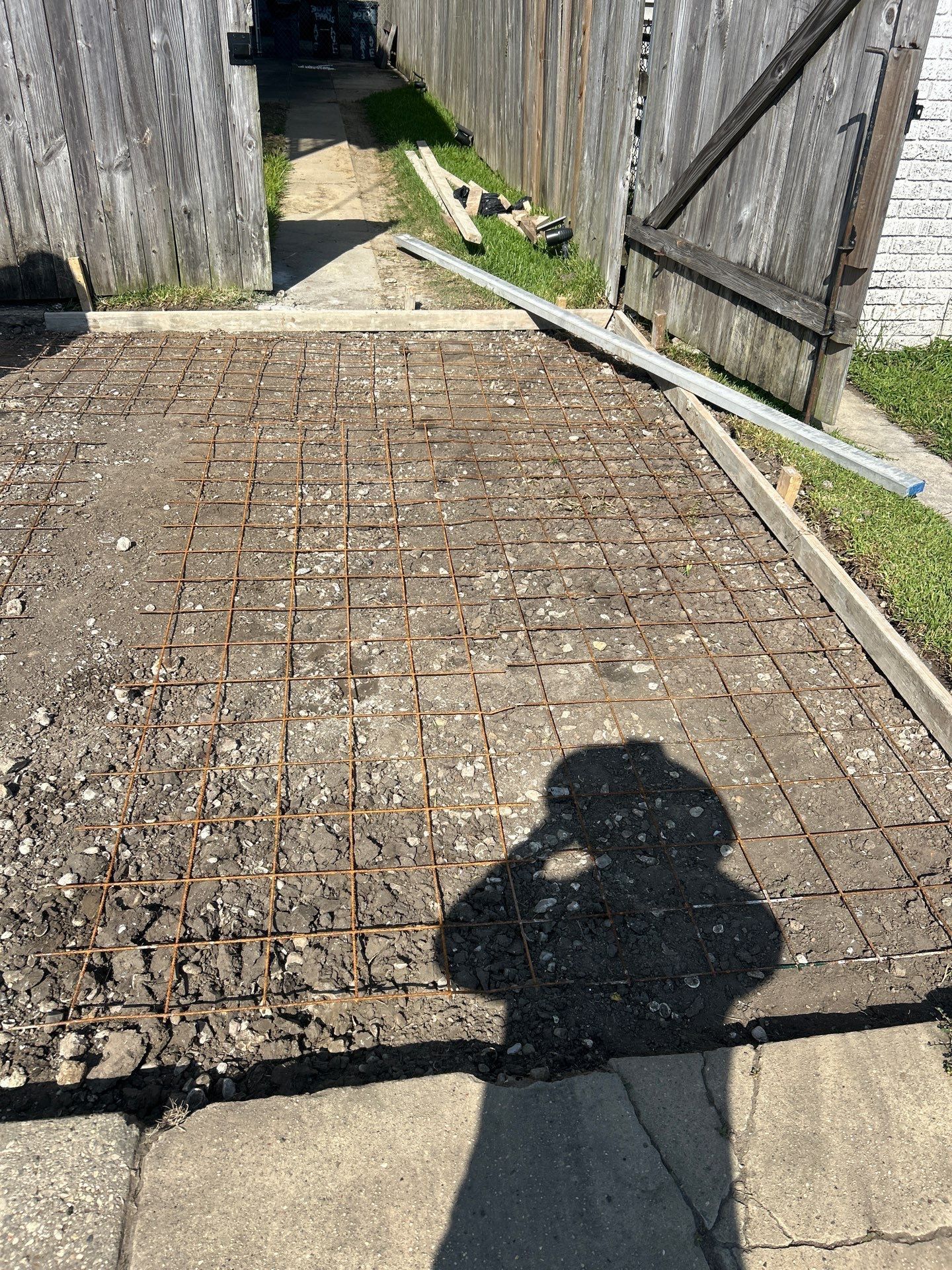 Shadow of a person over a driveway with exposed rebar grid, wooden gate, and concrete.