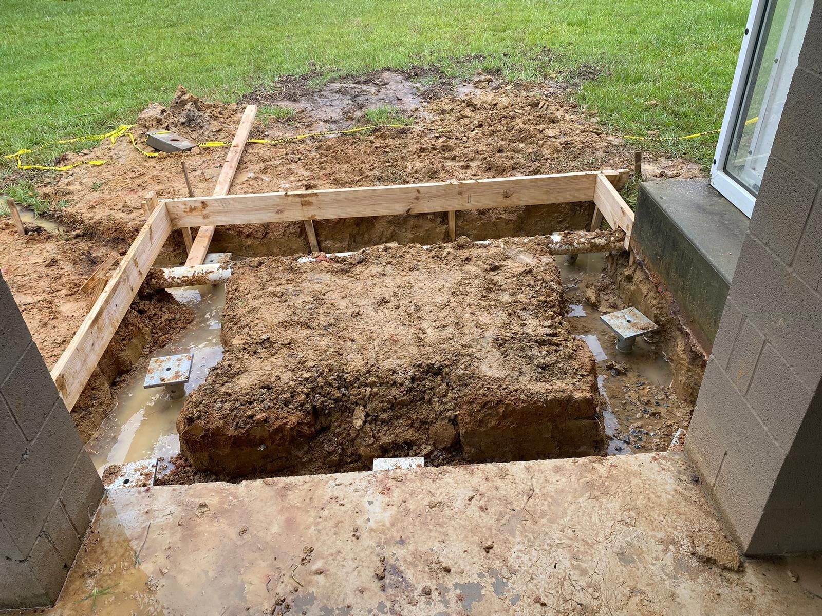 Construction site: wooden frame around a dirt foundation next to a building. Muddy ground and grass.