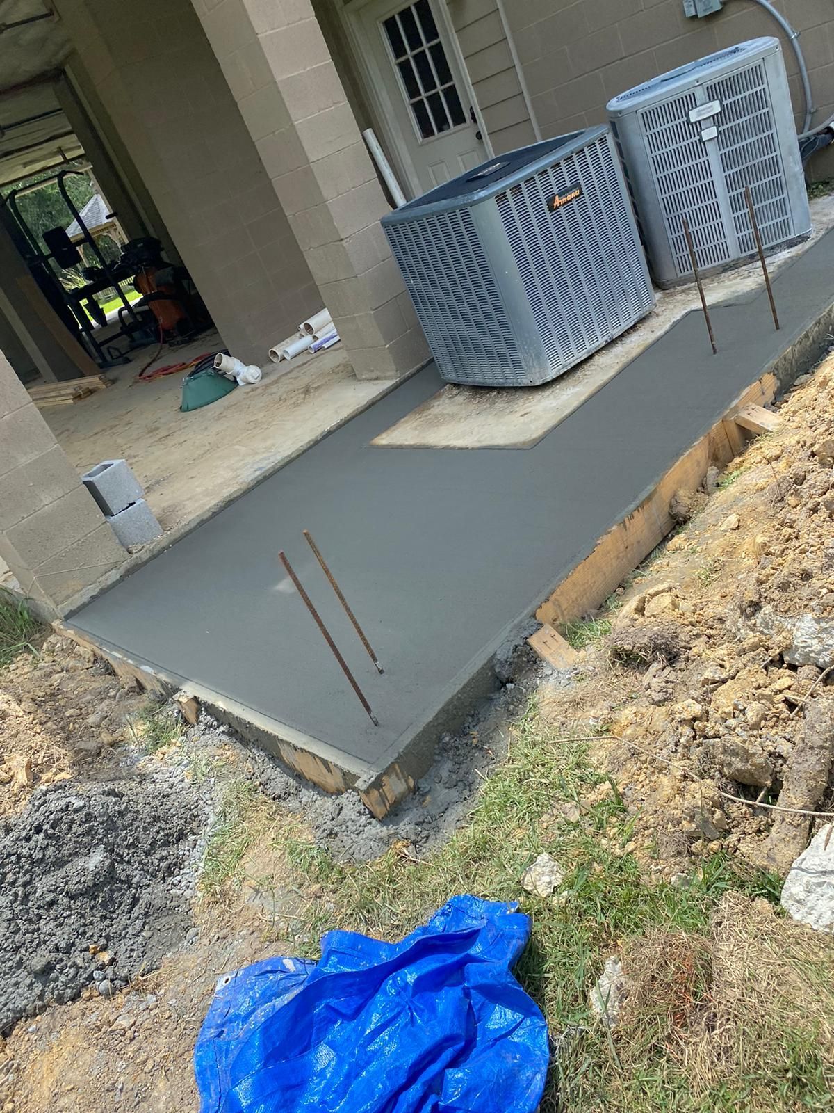 Freshly poured concrete walkway next to a building with two AC units. Wooden forms, dirt, and a blue tarp.