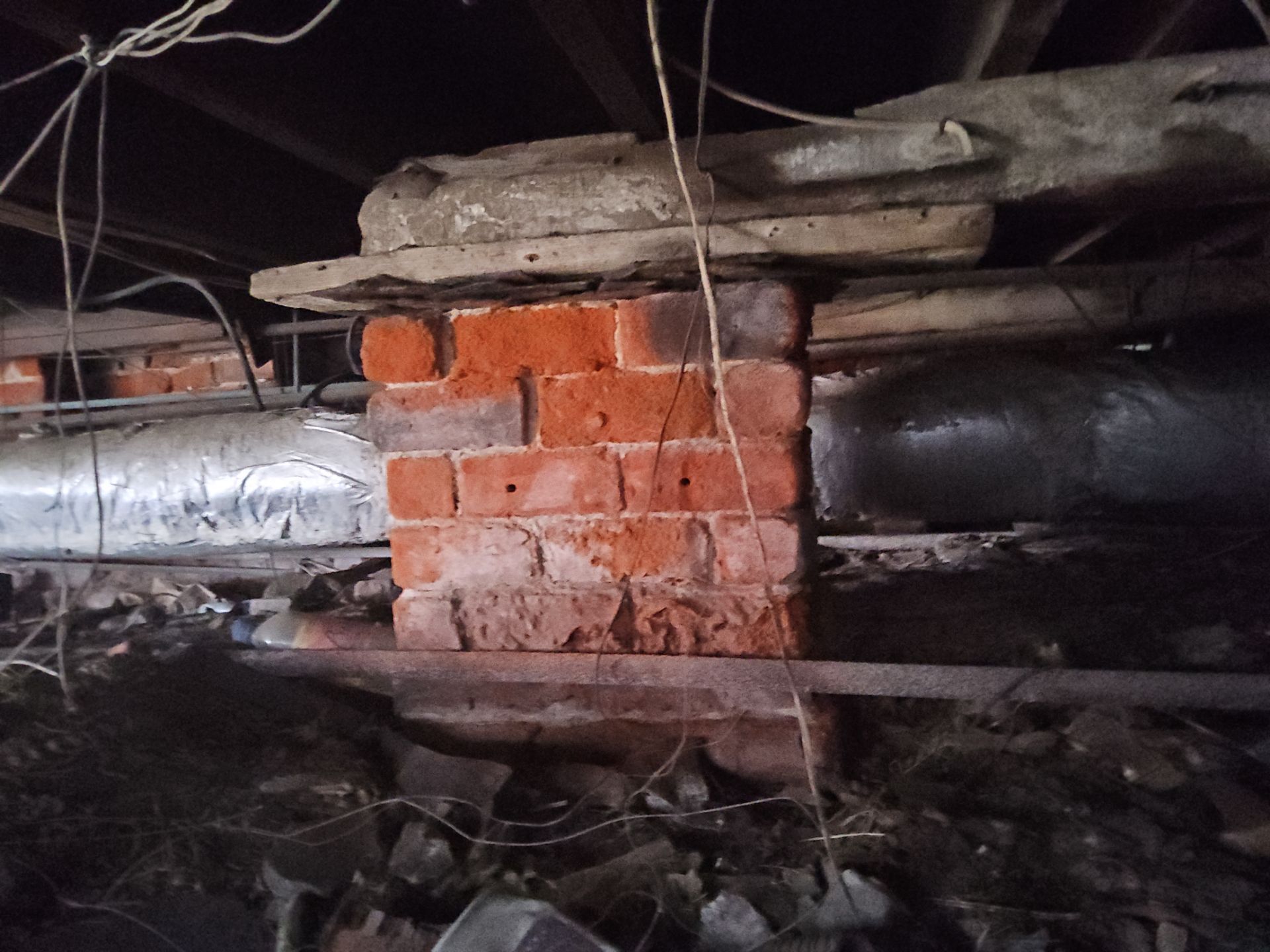 Brick support pillar in crawlspace, supporting wooden beam; red bricks, dark setting.
