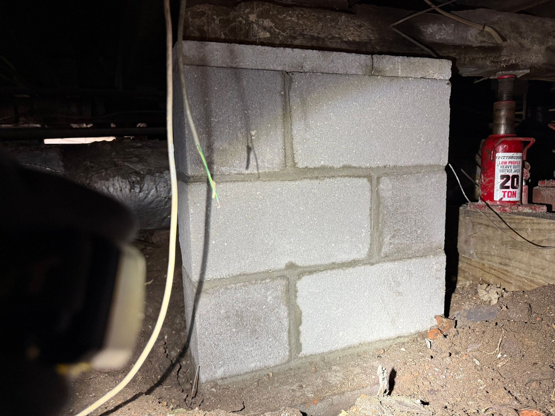 Gray cinder block pillar supporting a house, with a hydraulic jack visible behind it.