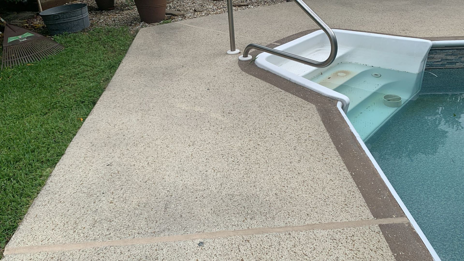 Concrete pool deck with pool steps and handrail; green grass at the left.