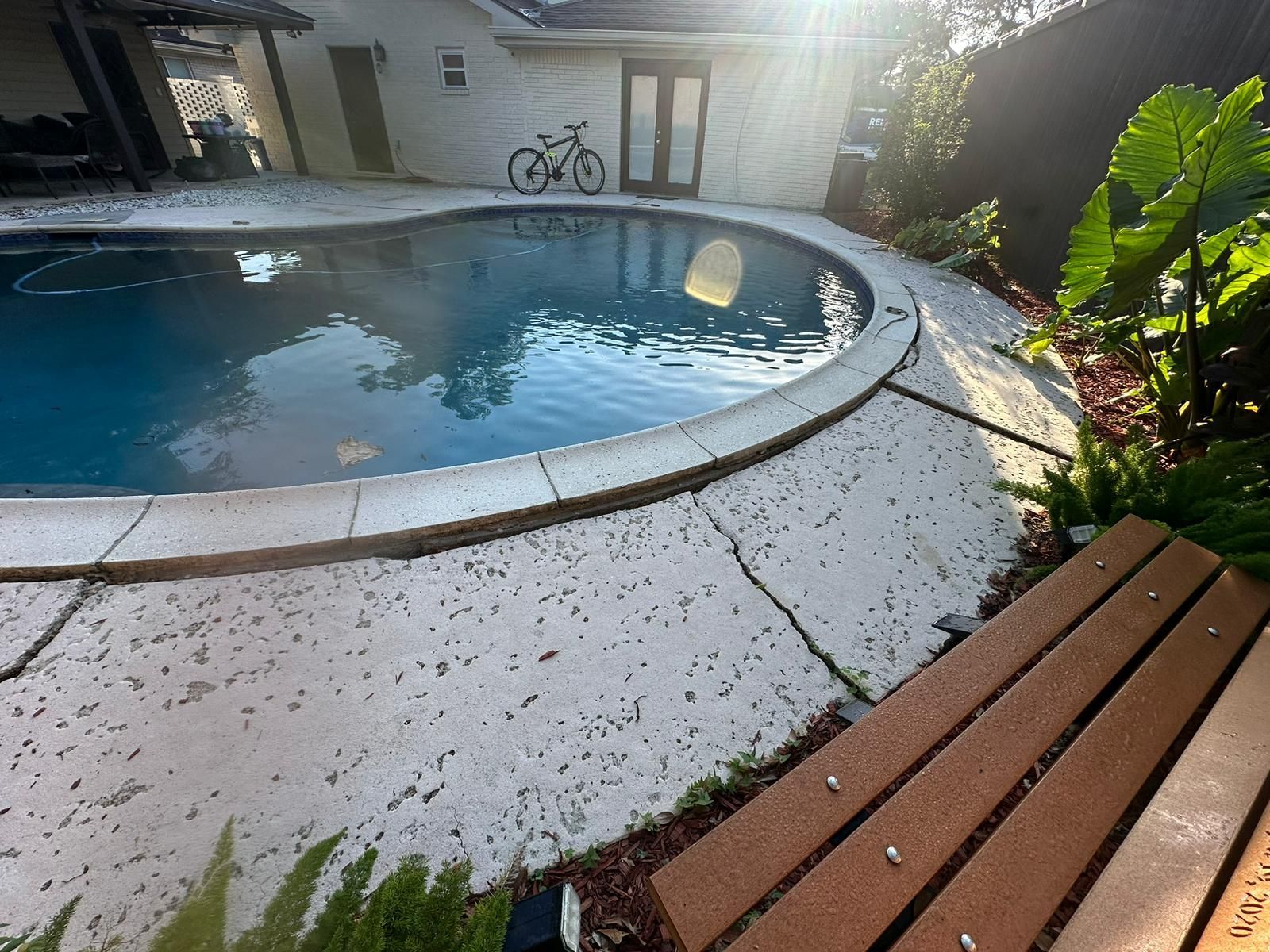 Pool with cracked concrete surround, bench, and bicycle near a house.