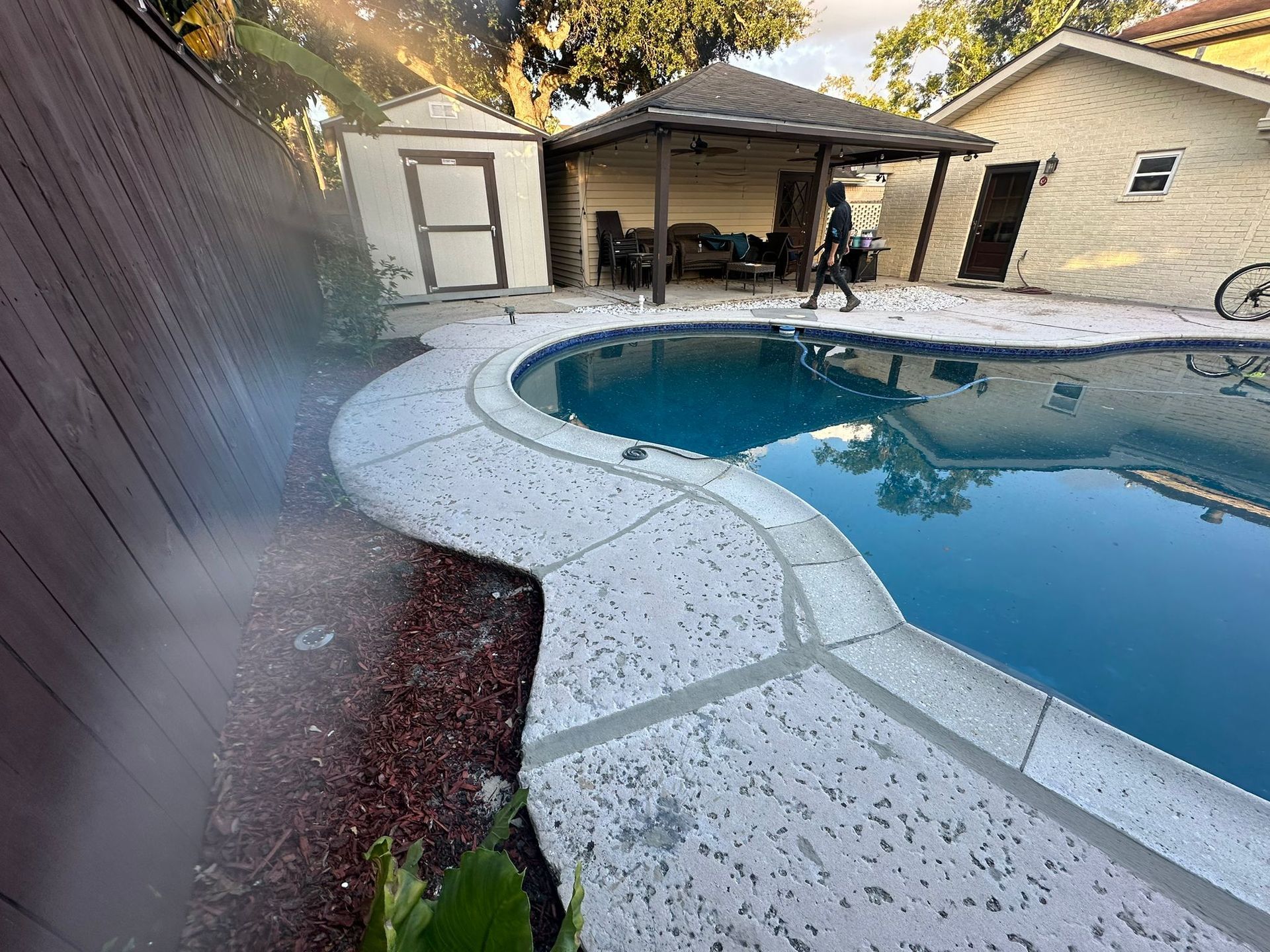 Poolside scene: Person walking near pool. Shed and covered patio in background.