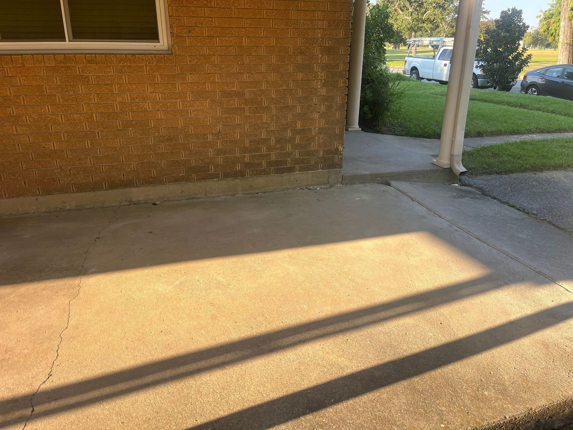 Concrete porch under a brick wall, with a shadow. Grass and cars are in the background.