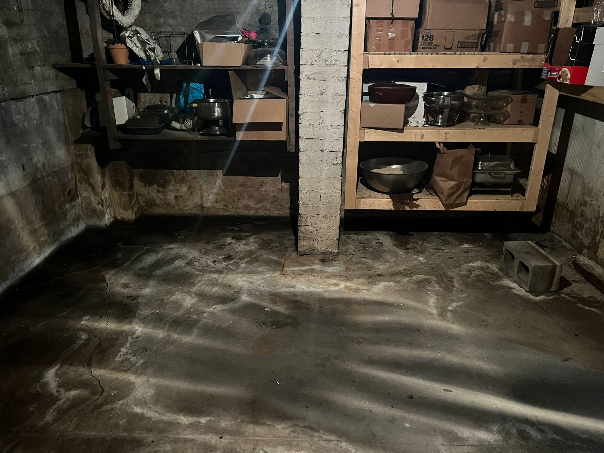 Basement interior with concrete floor, shelves, and a support beam; the floor shows water damage.