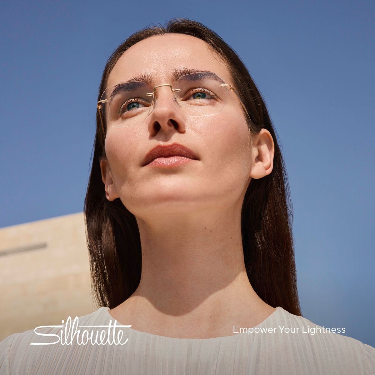 Woman wearing gold-rimmed glasses looks upwards, against a blue sky.