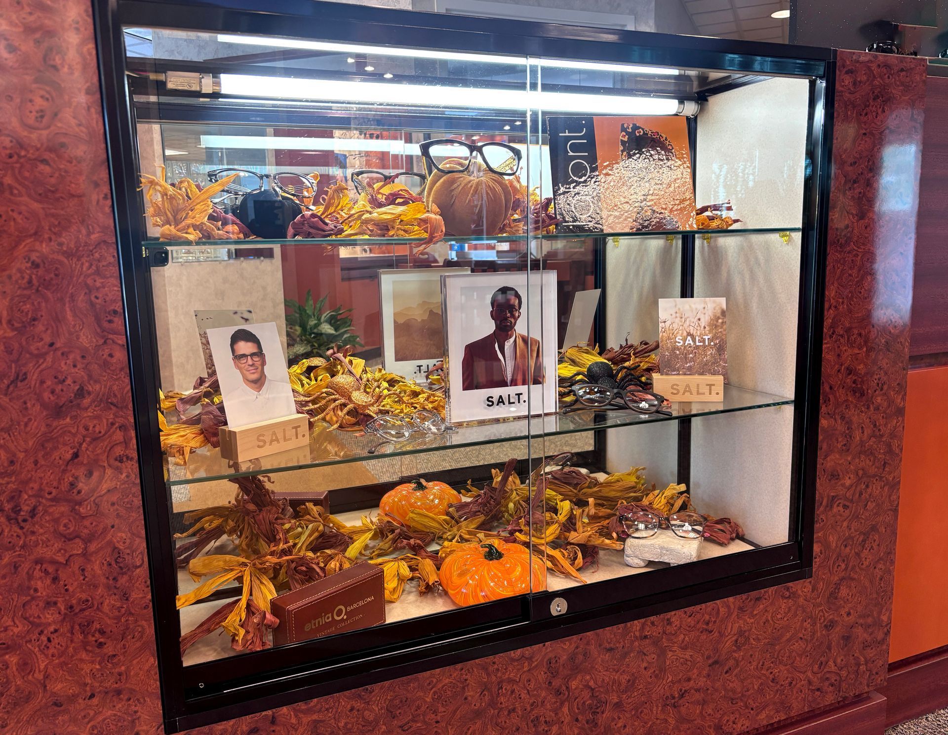 Glass memorial display with flowers, candles, and framed photos of three people on a red wall