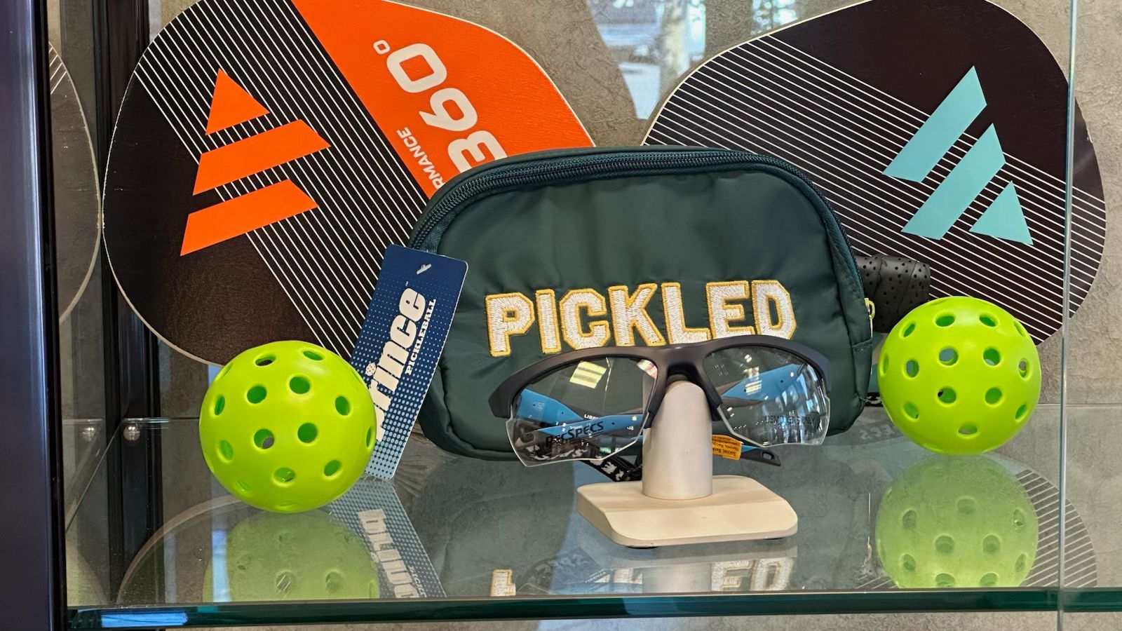 Pickleball paddles, balls, and a PICKLED bag on a glass shelf