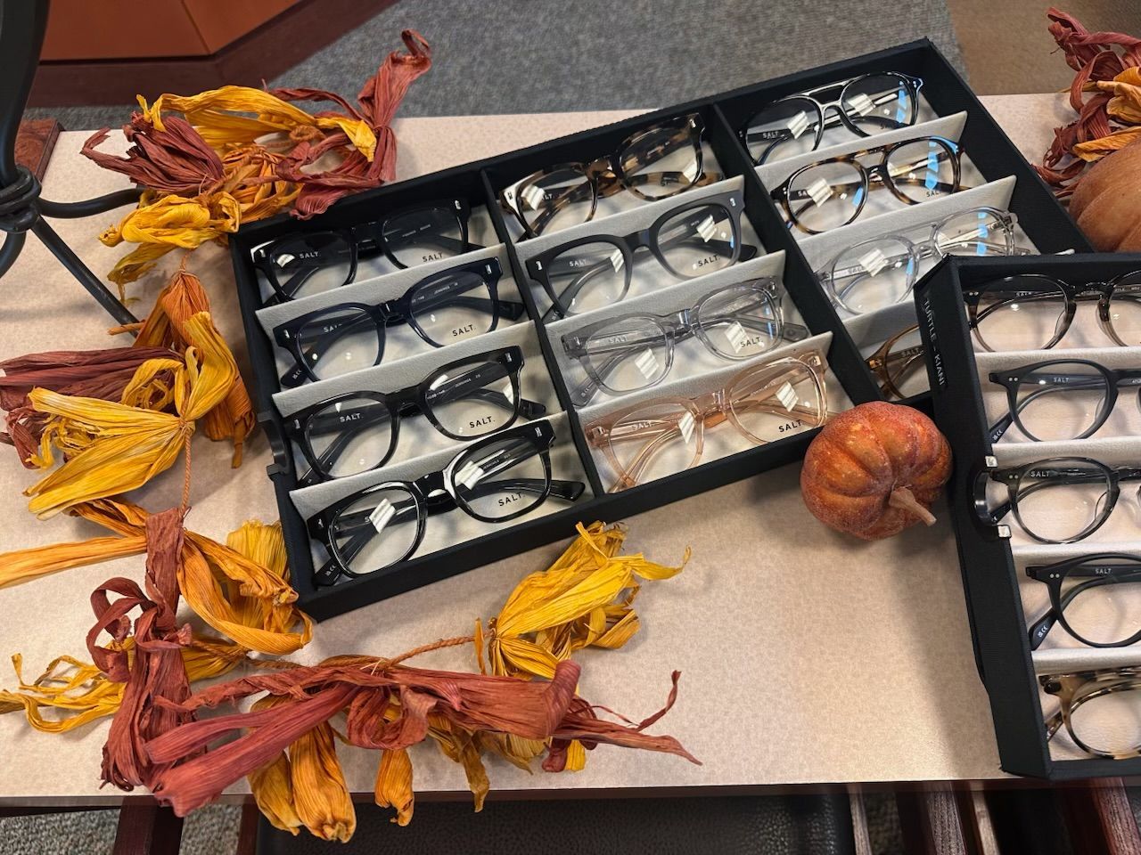 Eyeglasses in black trays with dried autumn leaves and a small pumpkin on a tabletop