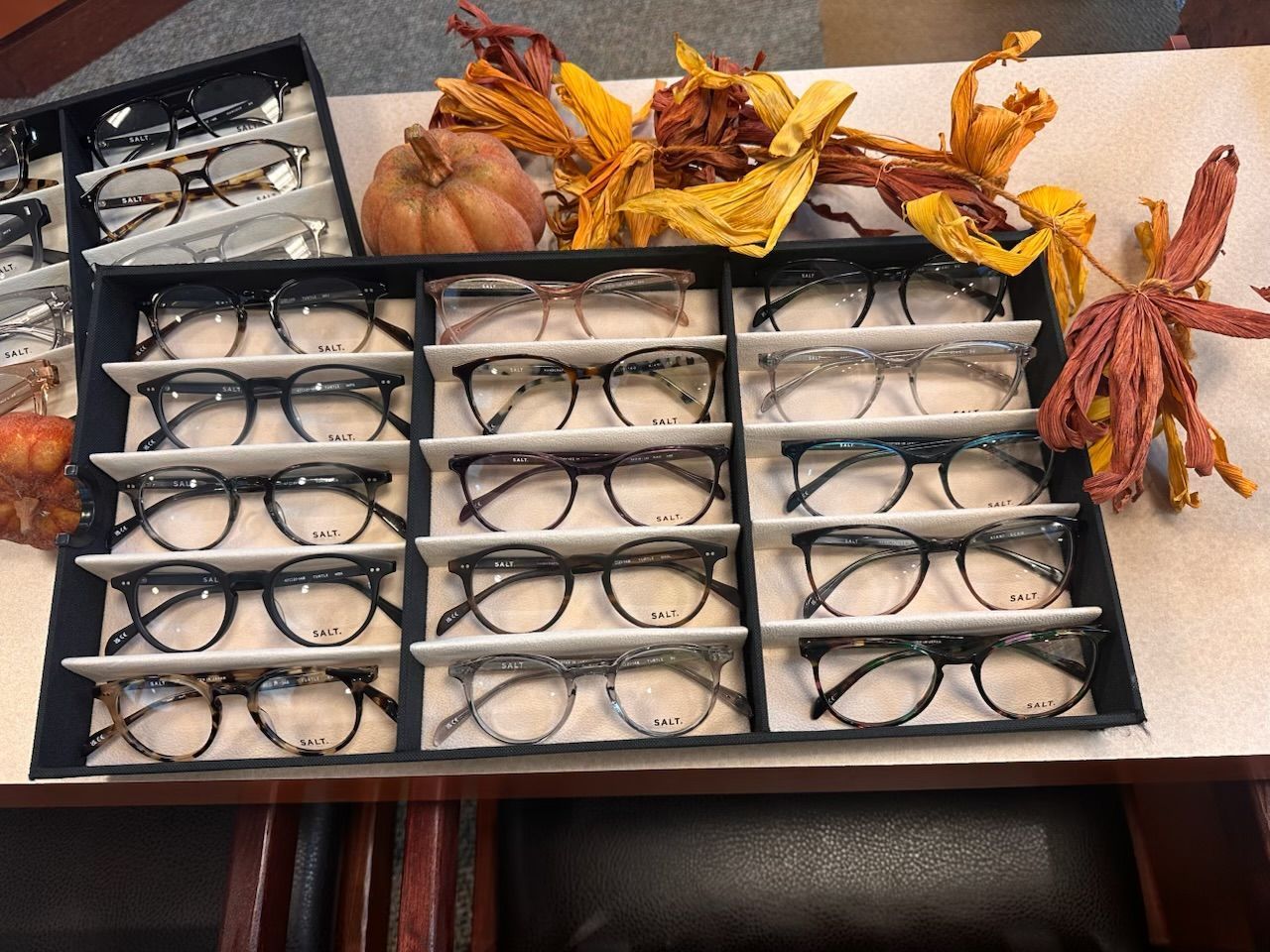 Neatly arranged eyeglasses in black trays on a table, with autumn decorations and leaves beside them