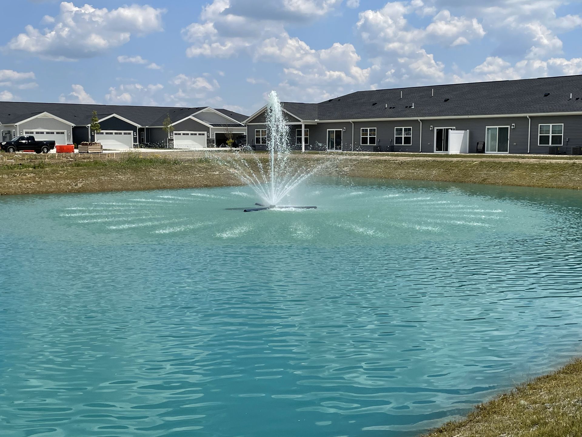 Pond with fountain spraying water, houses in background, blue sky, and green grass.