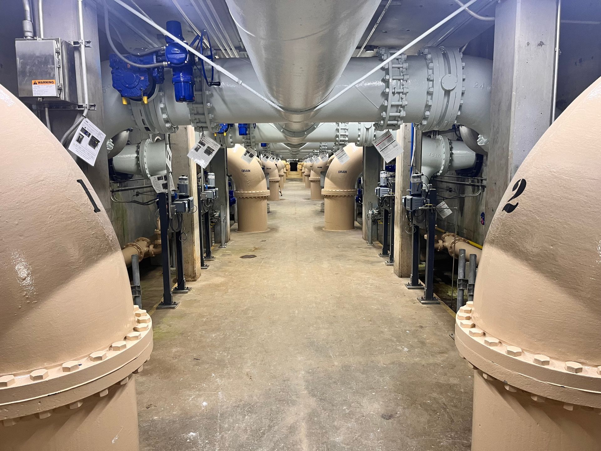 Interior view of a water treatment plant, beige pipes and equipment line a corridor with concrete floor.