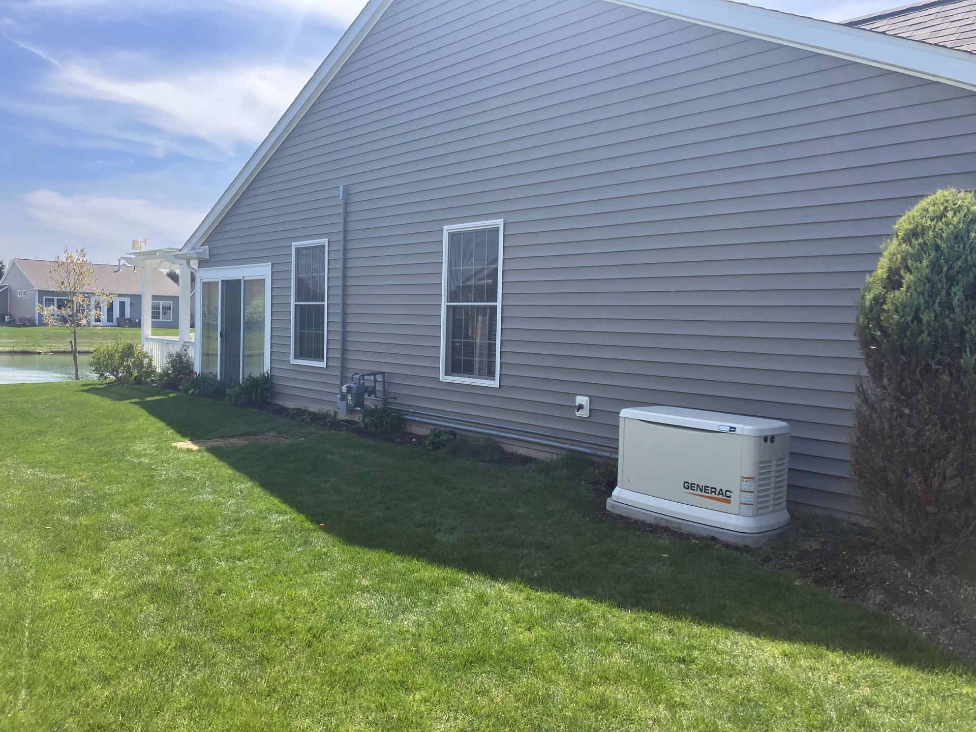 A gray house with a generator in the yard, next to a grassy area and a body of water.