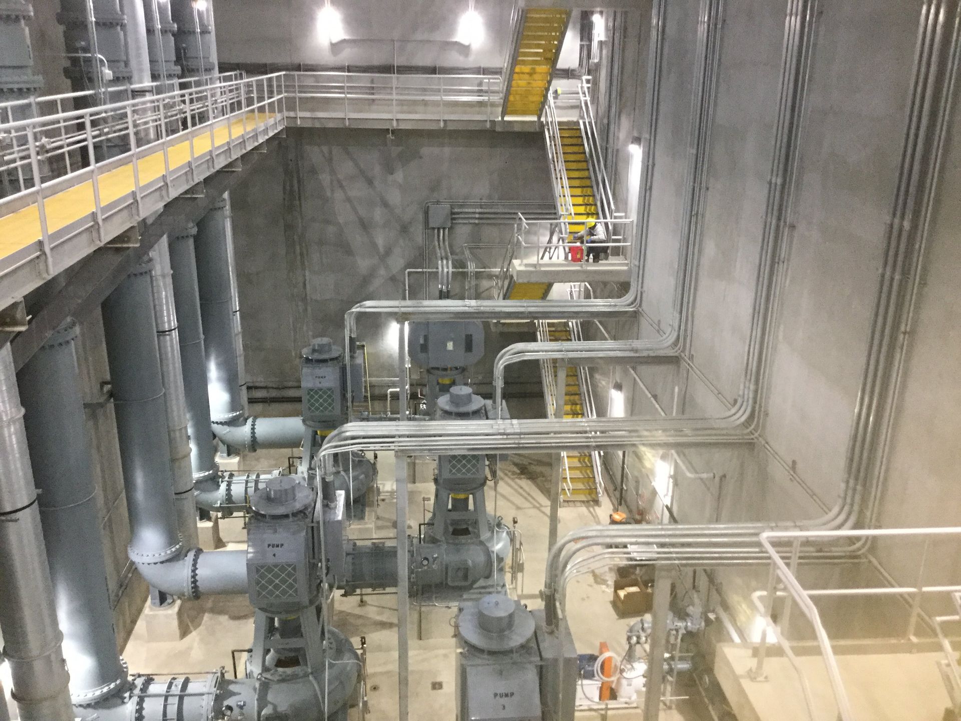 Pumps, pipes, and walkways in a concrete water treatment facility. Overhead electrical conduits and stairs are visible.