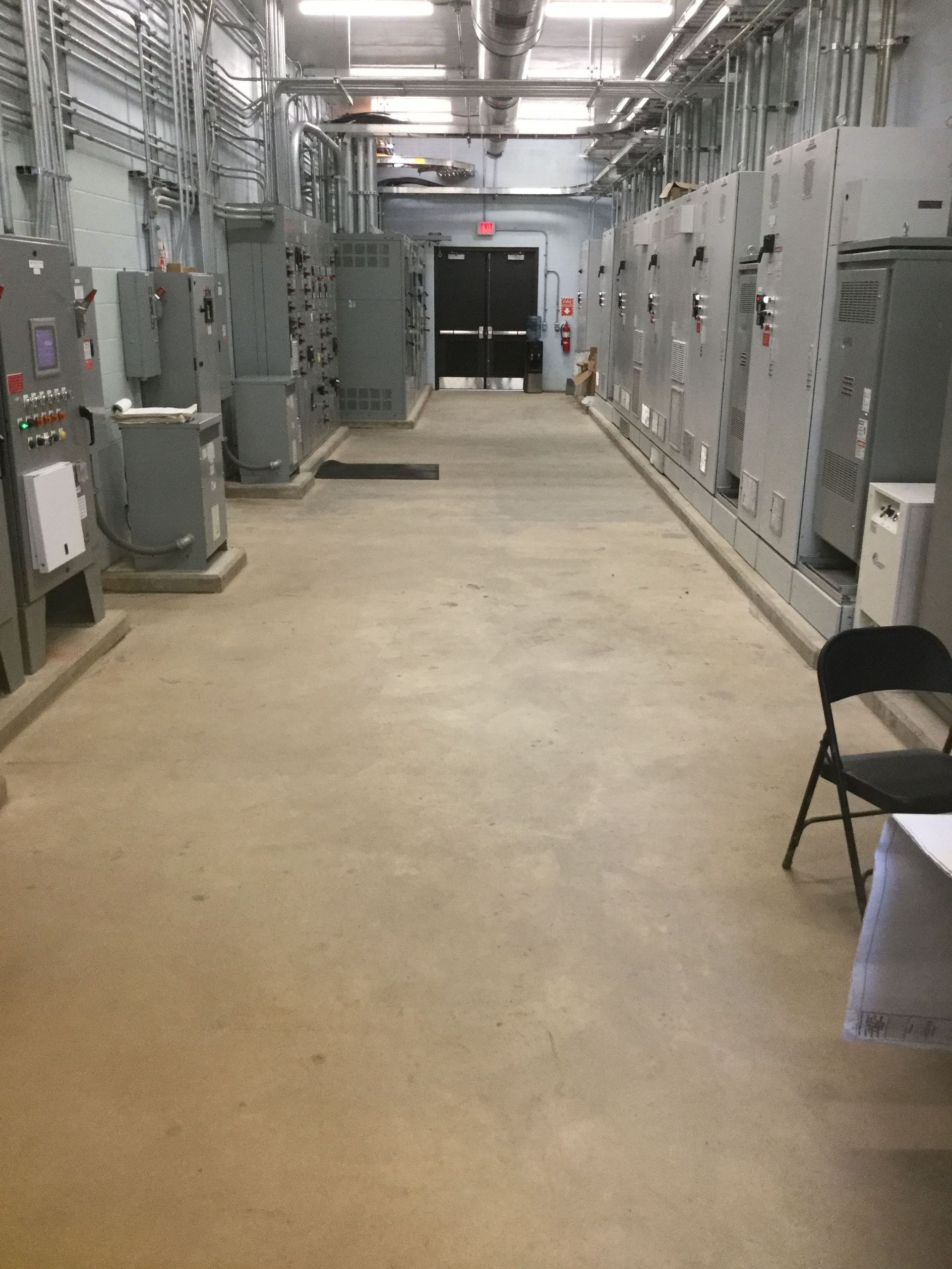 Electrical equipment room with concrete floor. Grey panels and conduit lines the walls. Black door at the end.