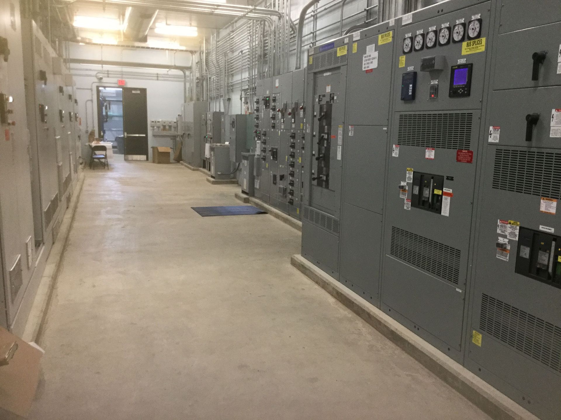 Electrical equipment room. Gray metal panels line the walls, concrete floor, overhead lighting, and a doorway at the end of the hallway.