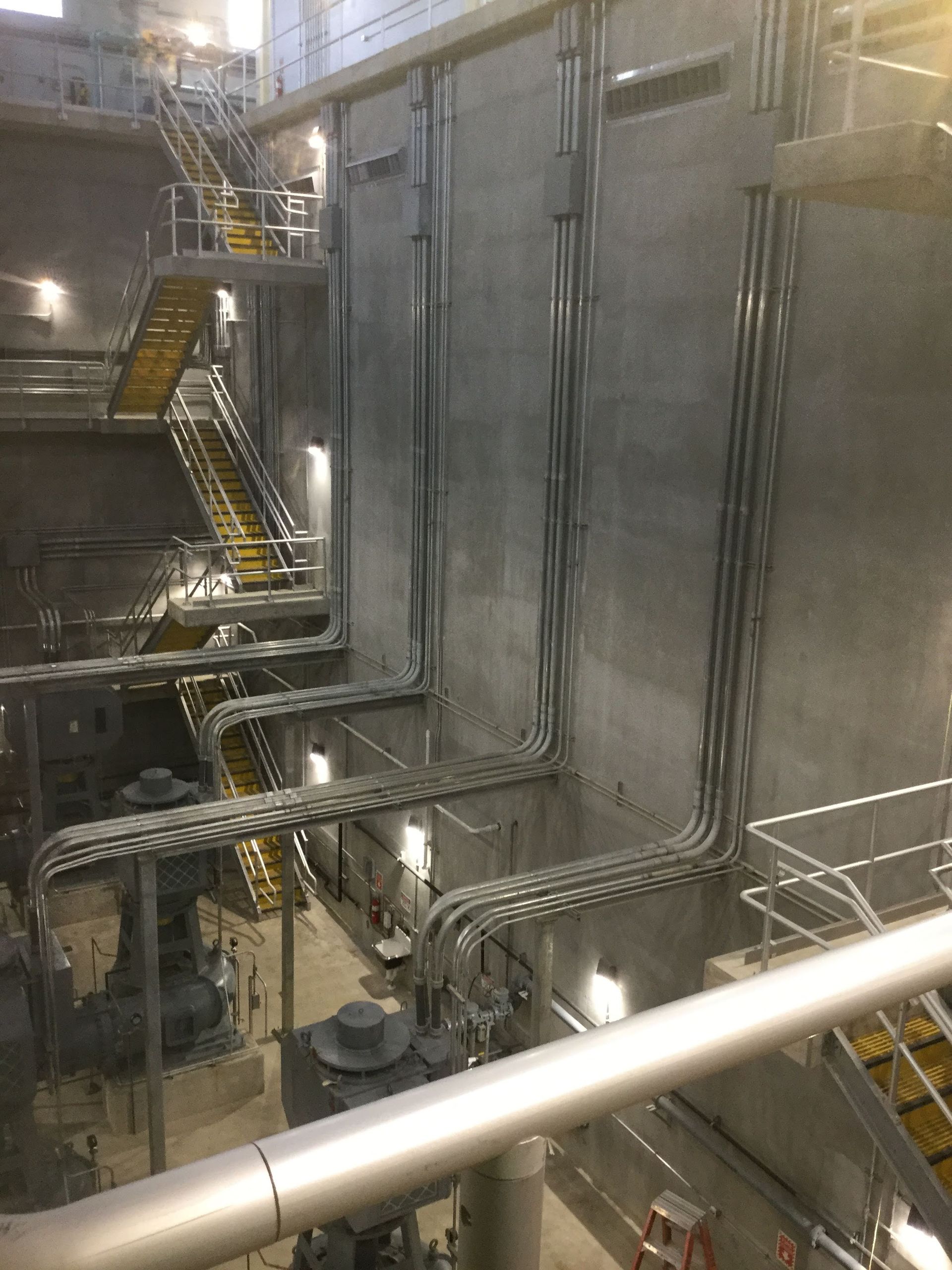 Interior of a concrete industrial space with metal piping and staircases. Yellow steps, grey walls.