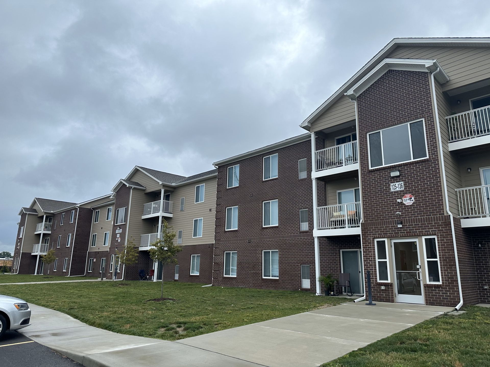 Apartment building with brown brick and tan siding on a cloudy day; green lawn and sidewalk in front.