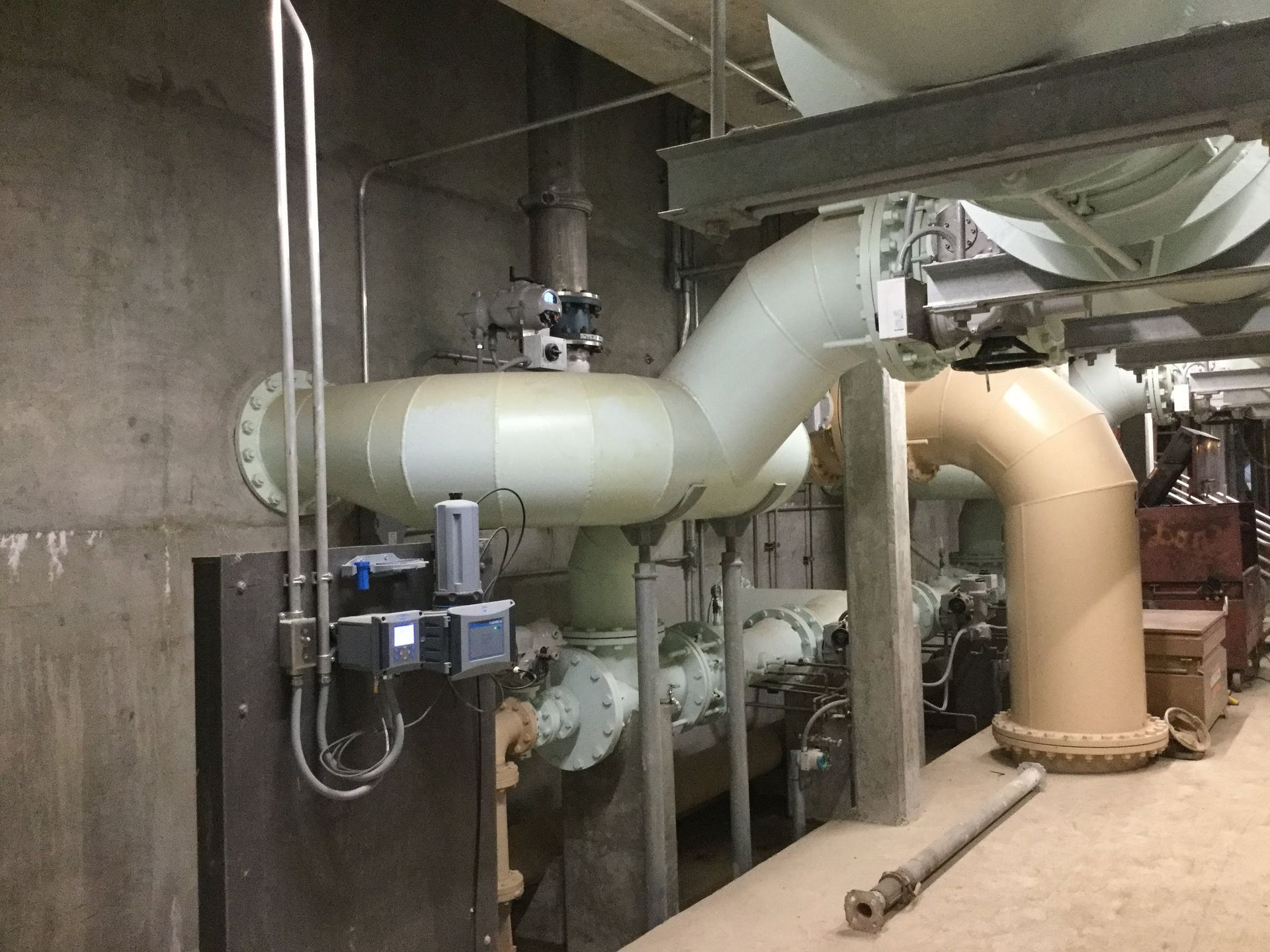 Industrial piping in a concrete room; pipes are beige and off-white. A control panel is mounted on a wall.
