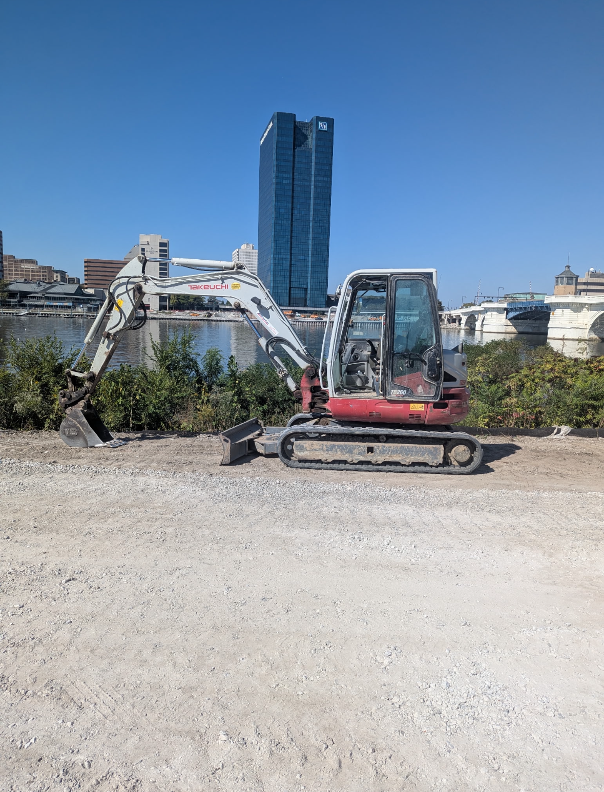 Small excavator on gravel, with tall, blue glass building and sky in background.