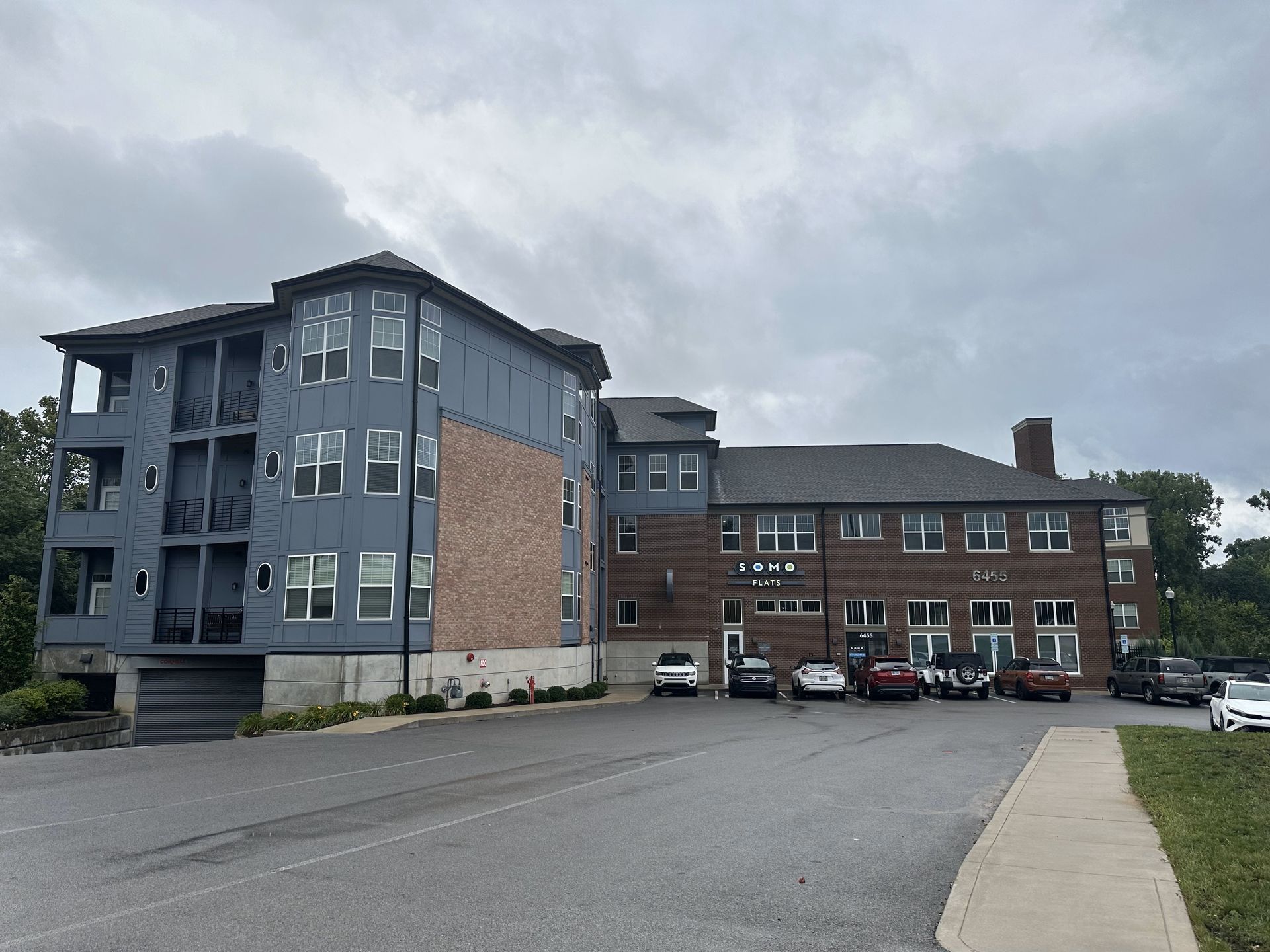 A multi-story building with gray, brick, and blue siding, a parking lot, and a cloudy sky.