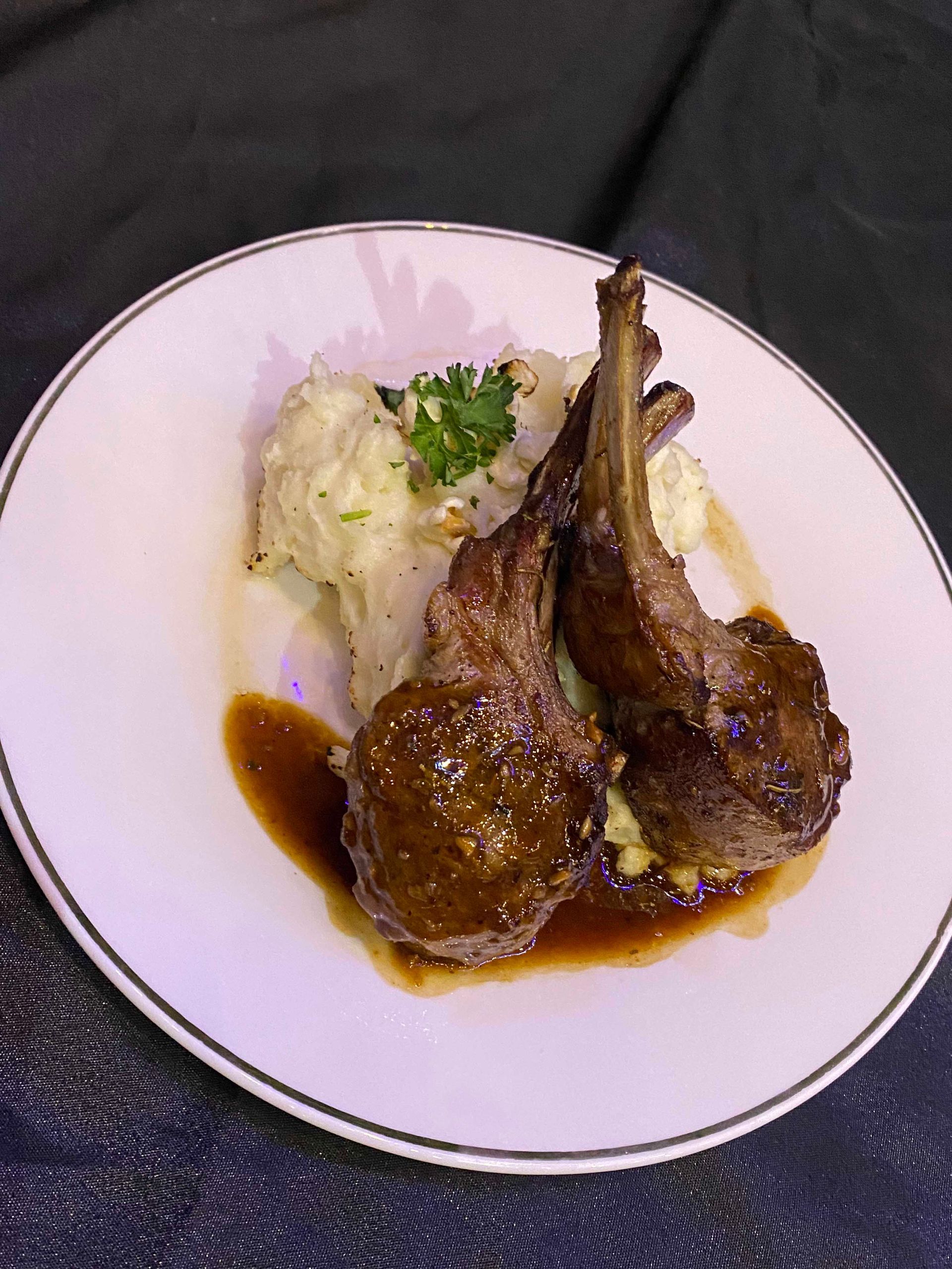 Two lamb chops on mashed potatoes, drizzled with sauce, on a white plate.