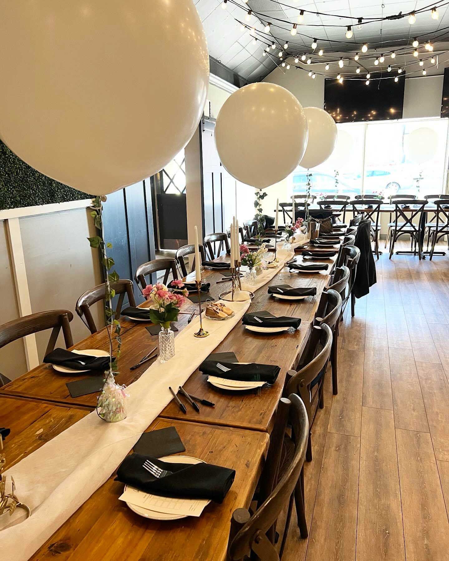 Long wooden table set for a meal in a restaurant with white balloons and string lights.