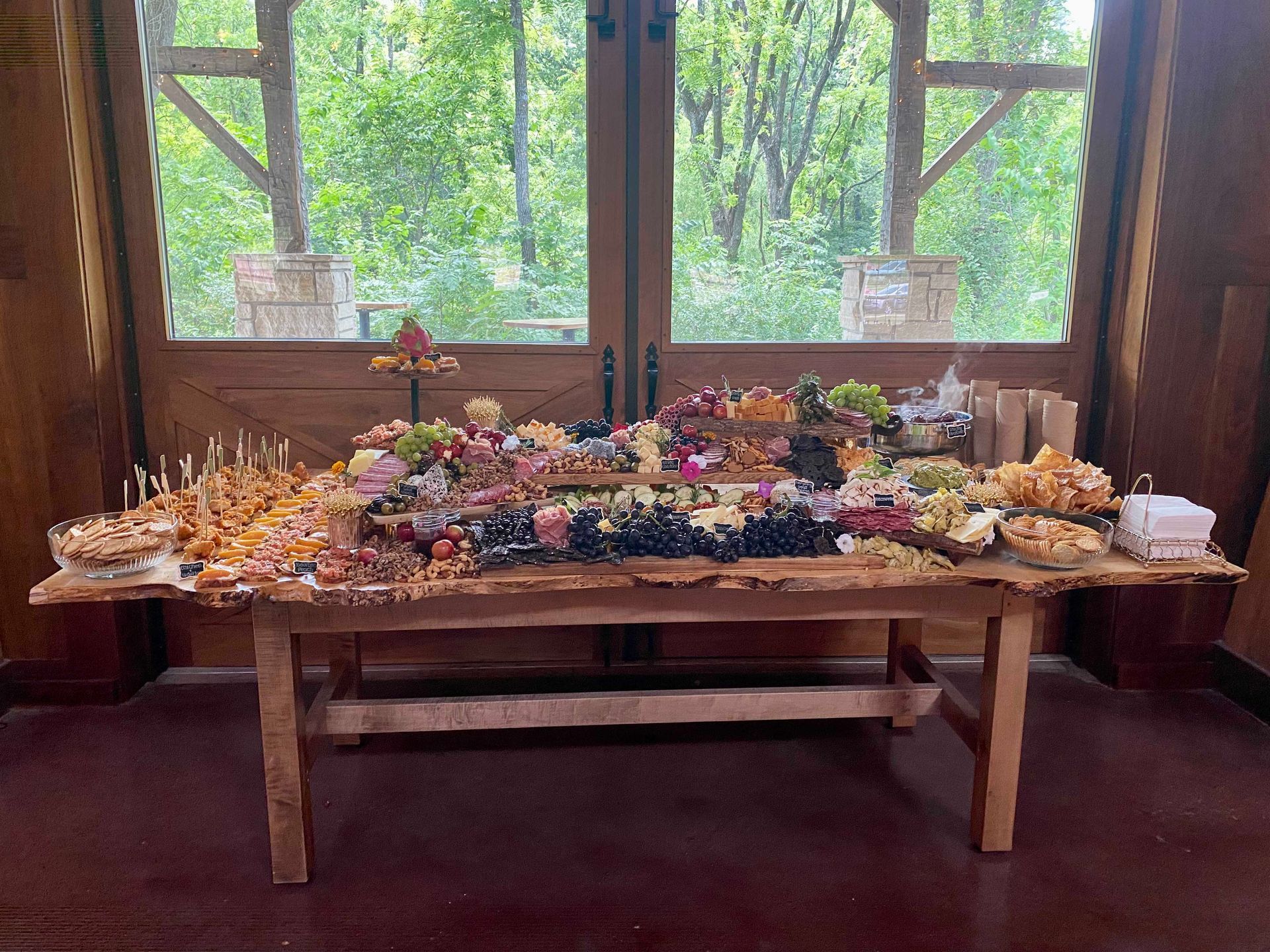 A wooden table laden with a charcuterie spread, in front of large windows overlooking a forest.