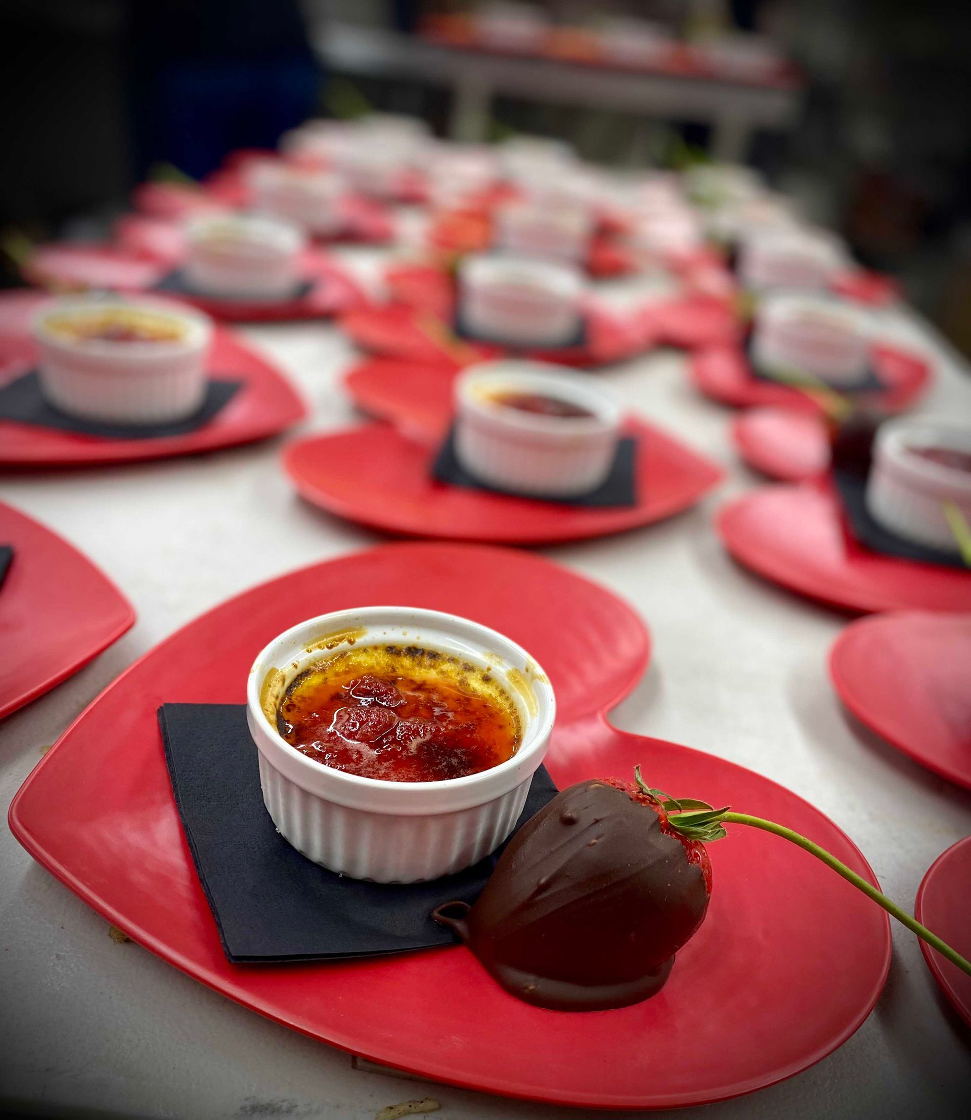 Desserts on heart-shaped red plates: crème brûlée and chocolate-covered strawberries, arranged in rows.