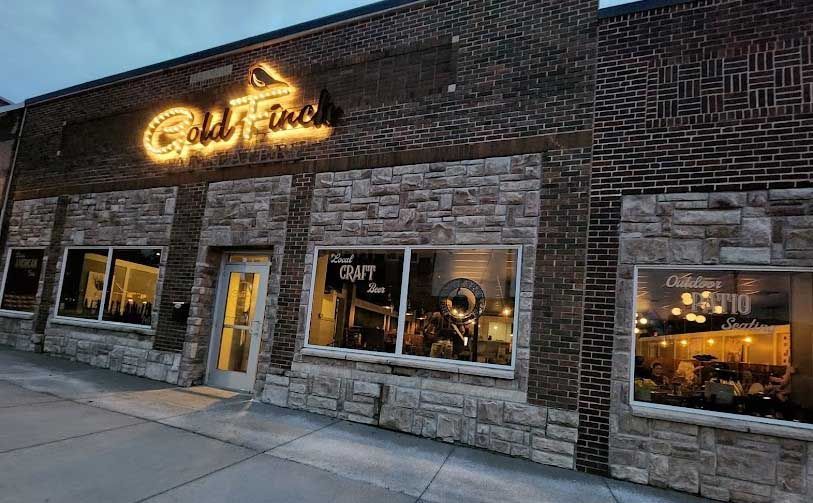 Exterior of Gold Finch restaurant with a neon sign above the entrance, brick and stone facade, windows with interior views.