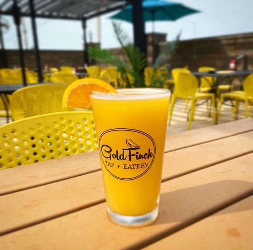 Glass of orange juice with orange slice at Goldfinch Cafe + Eatery outdoor patio, yellow chairs, sunny day.