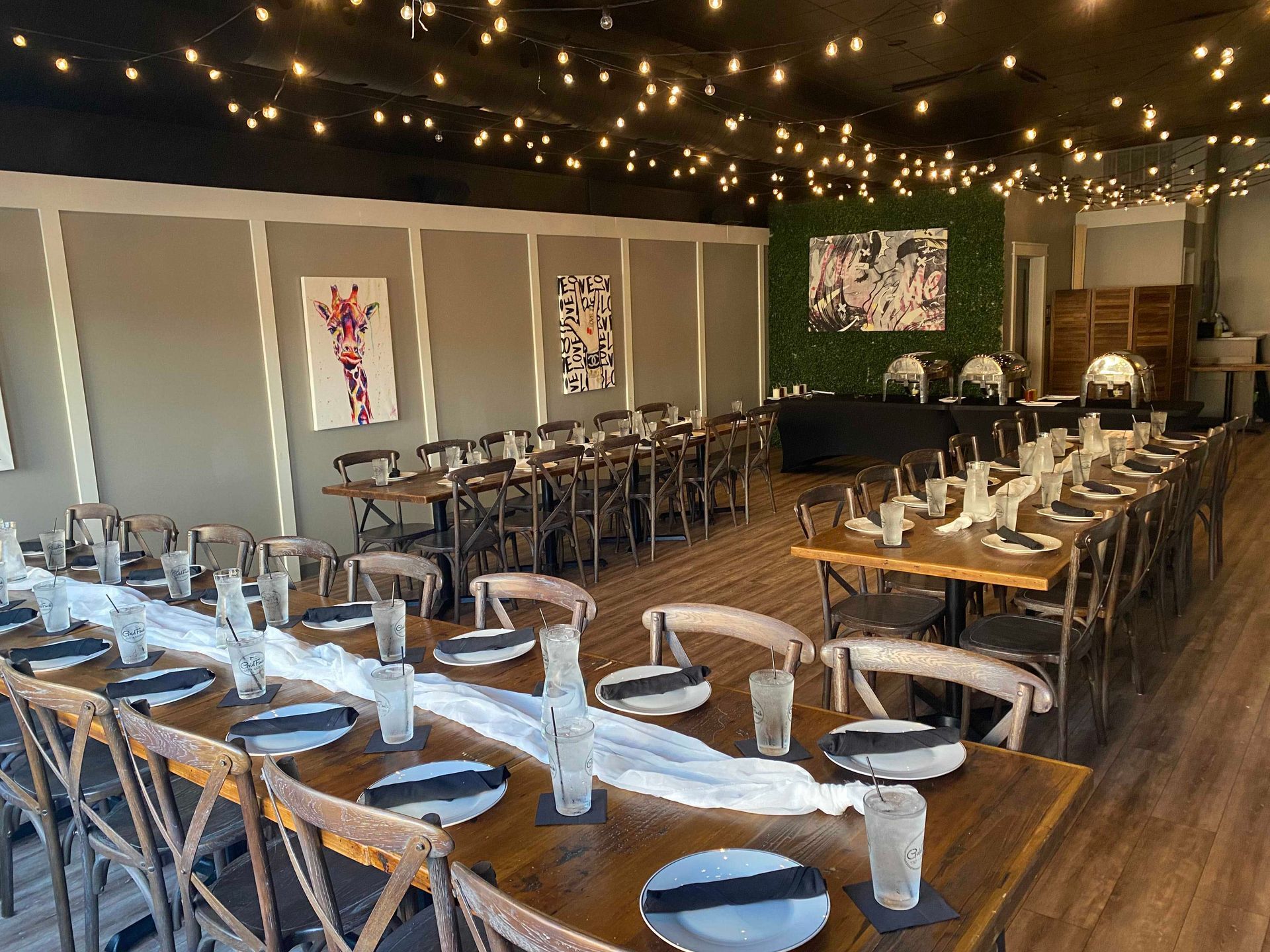 Dining room set up for an event, long wooden tables with place settings, string lights overhead.