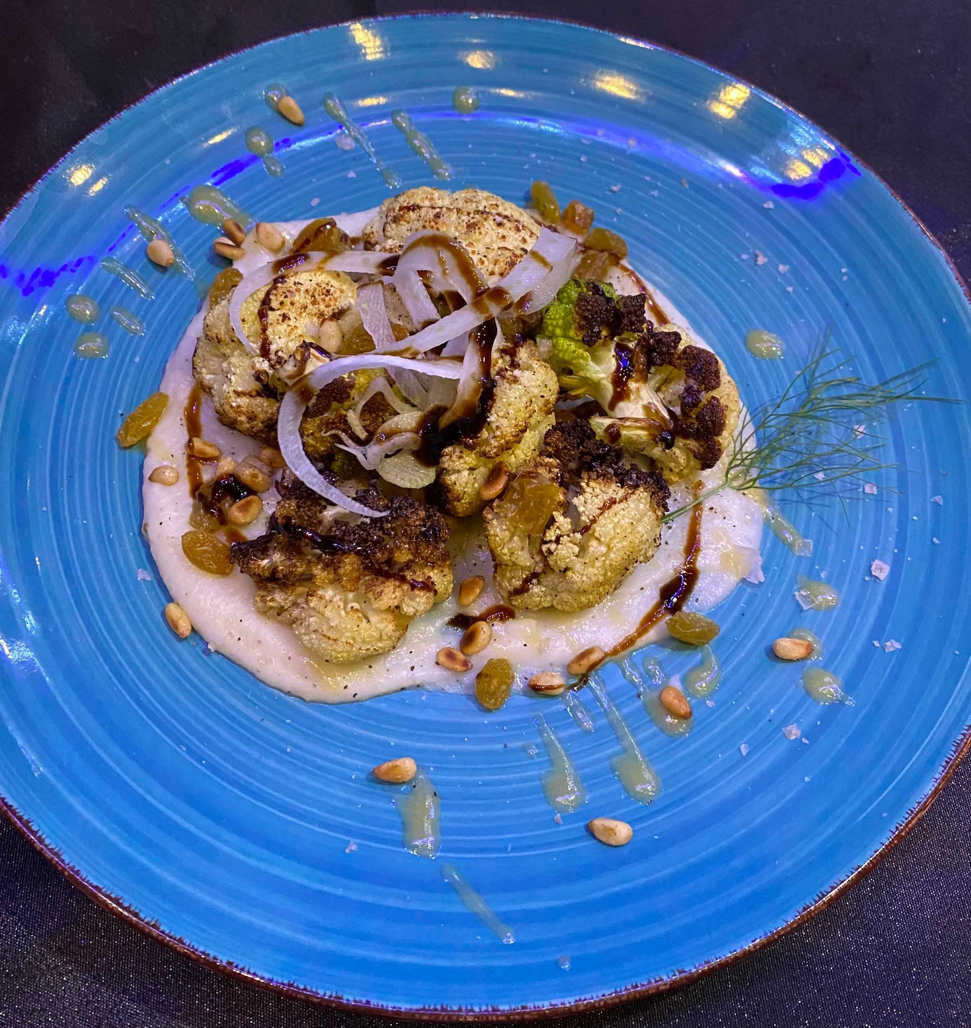 Roasted cauliflower on creamy white base, drizzled with sauce and sprinkled with raisins and pine nuts on a blue plate.