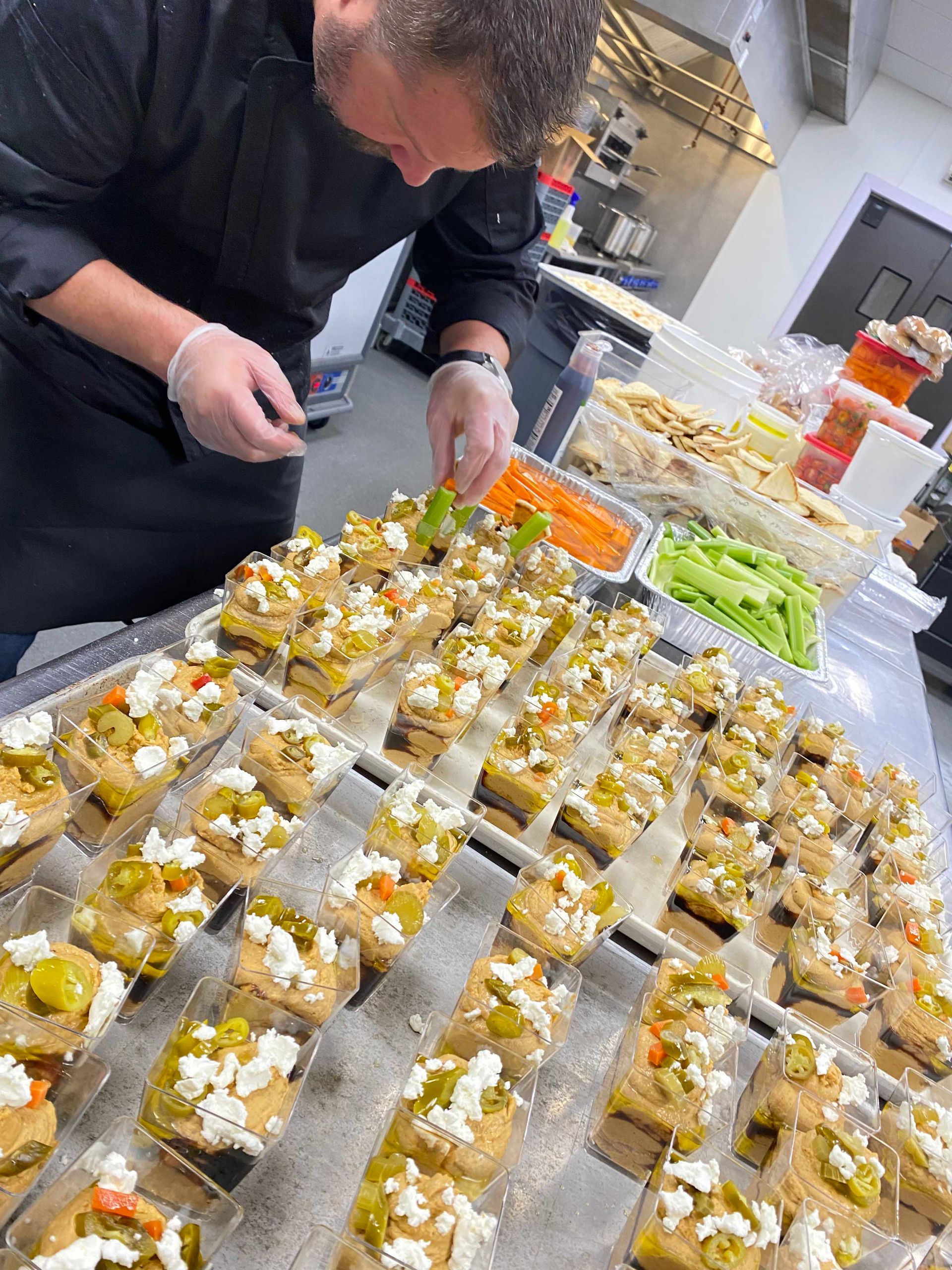 Chef garnishing hors d'oeuvres arranged on trays. Kitchen setting, various toppings, gloves, black chef's coat.
