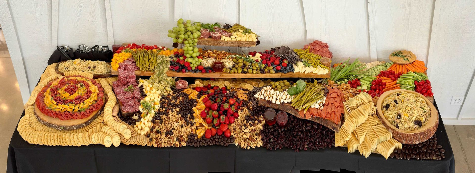 A large food display with various items. Pasta, fruit, vegetables are arranged on a dark table, against a white wooden backdrop.