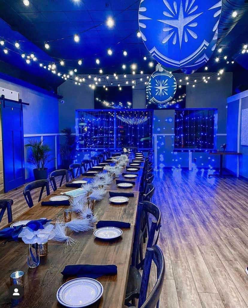 Long wooden table set for a formal gathering, decorated with blue accents. Room bathed in blue light.