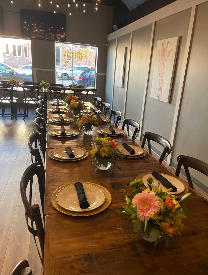 Long wooden table set for a meal in a restaurant with floral centerpieces and plates.