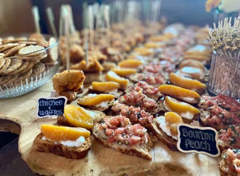Appetizer spread on a wooden board: chicken skewers, bruschetta with peaches, crackers, and signs.
