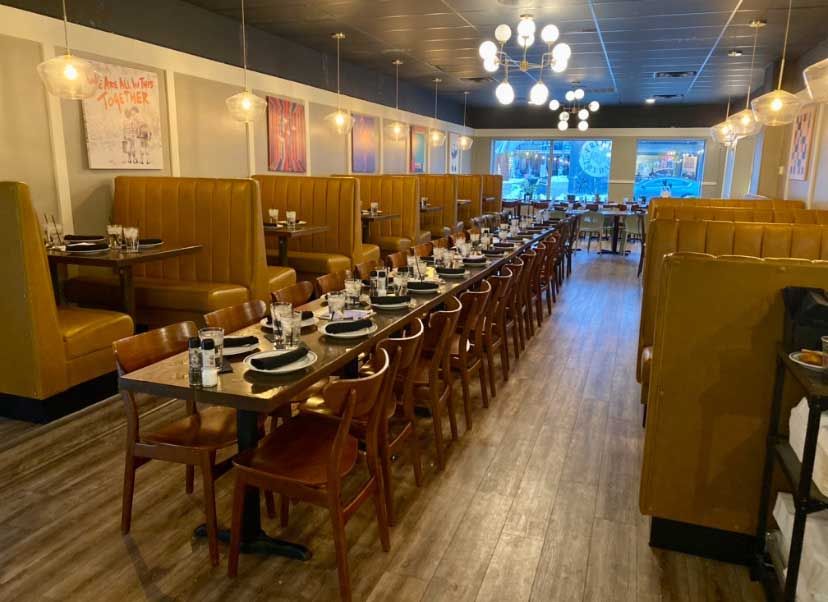 Restaurant interior with long table set for a group, booths with mustard-colored upholstery, overhead globe lights.