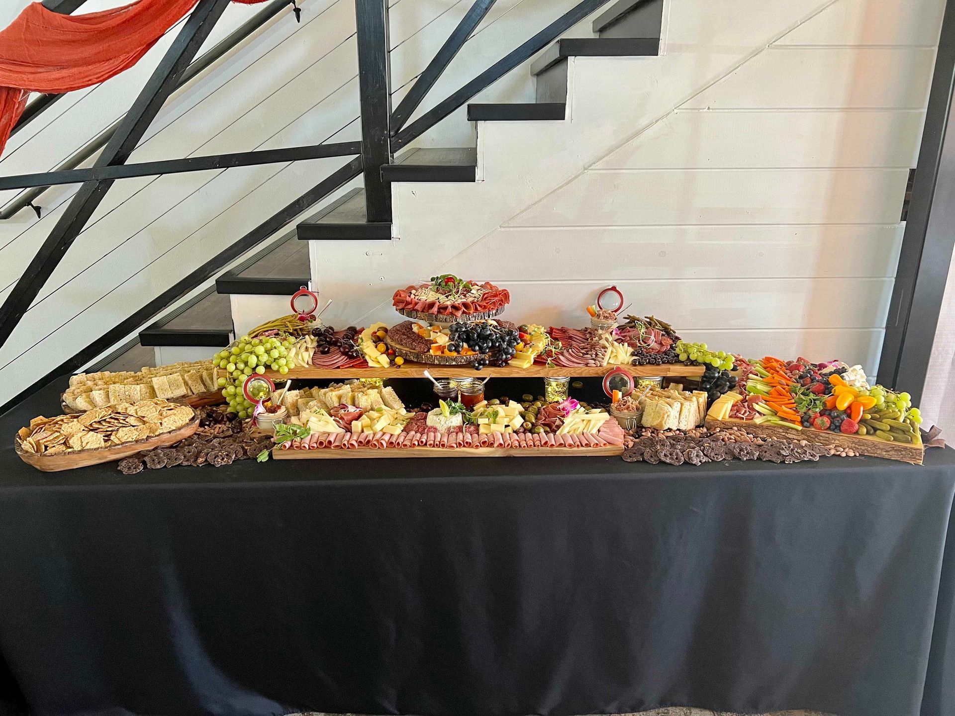 Charcuterie spread on a black-covered table with fruit, cheese, and crackers, set against a staircase.