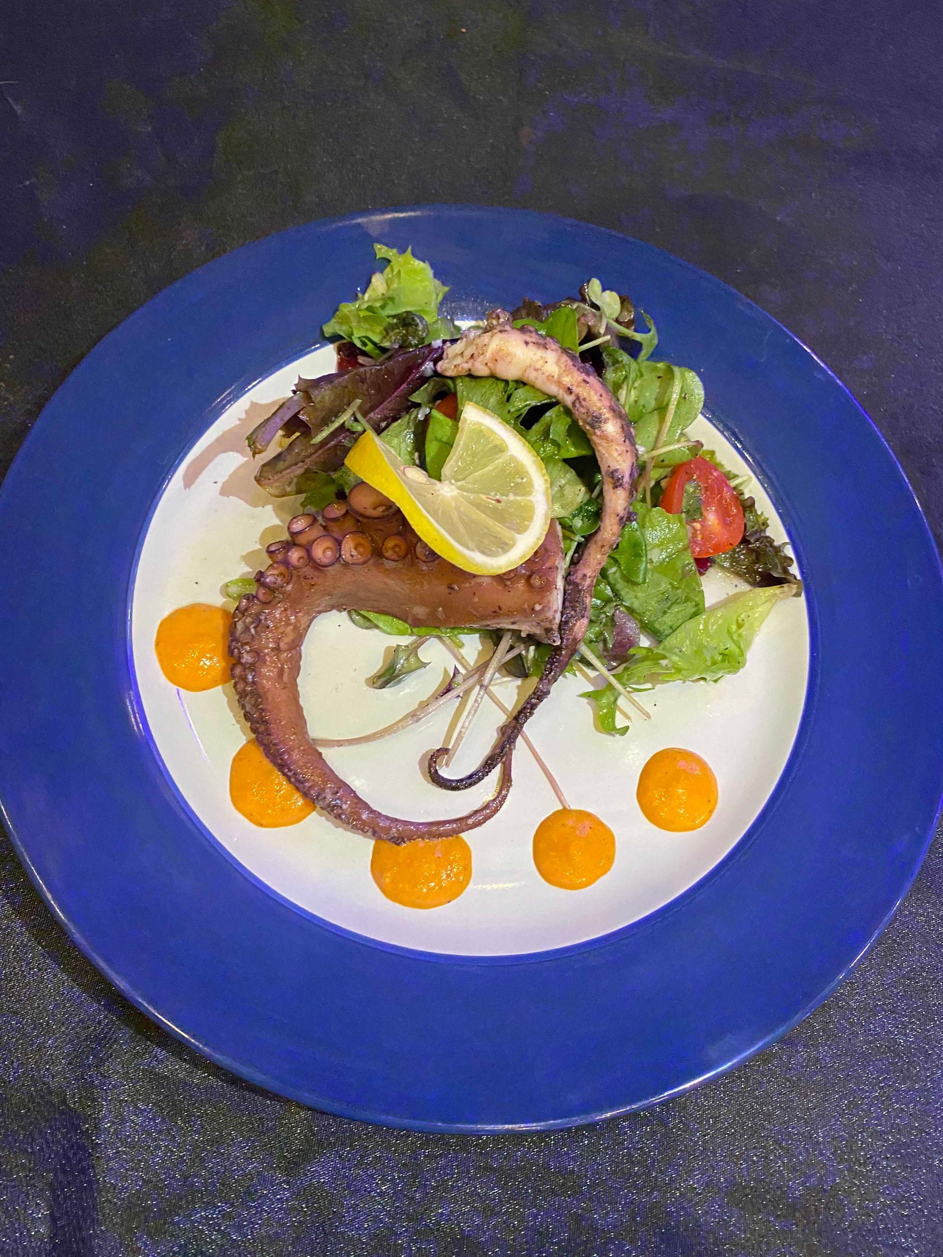 Grilled octopus with salad, lemon, and orange sauce on a blue-rimmed plate.