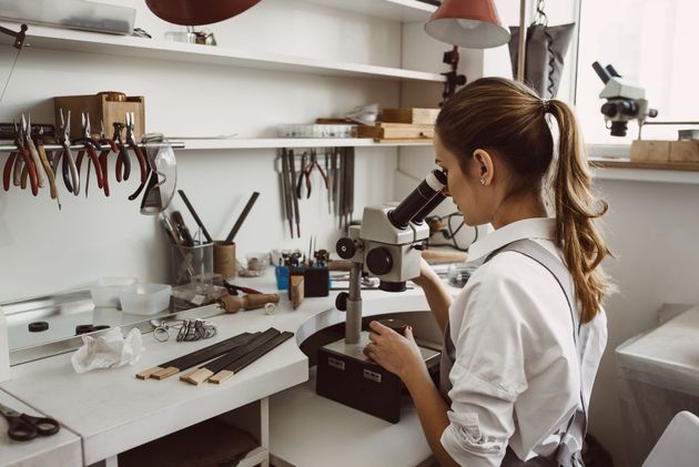 Person using a microscope at a workbench in a bright jewelry or craft workshop