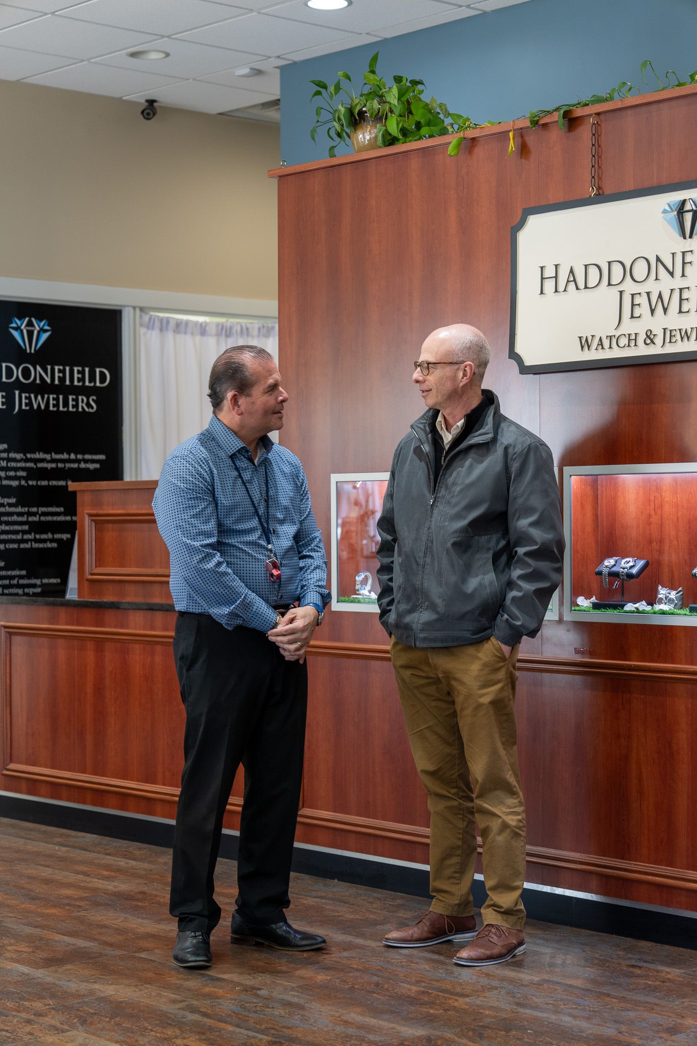 Two men talking in a jewelry store, standing by a glass display case and wood counter.