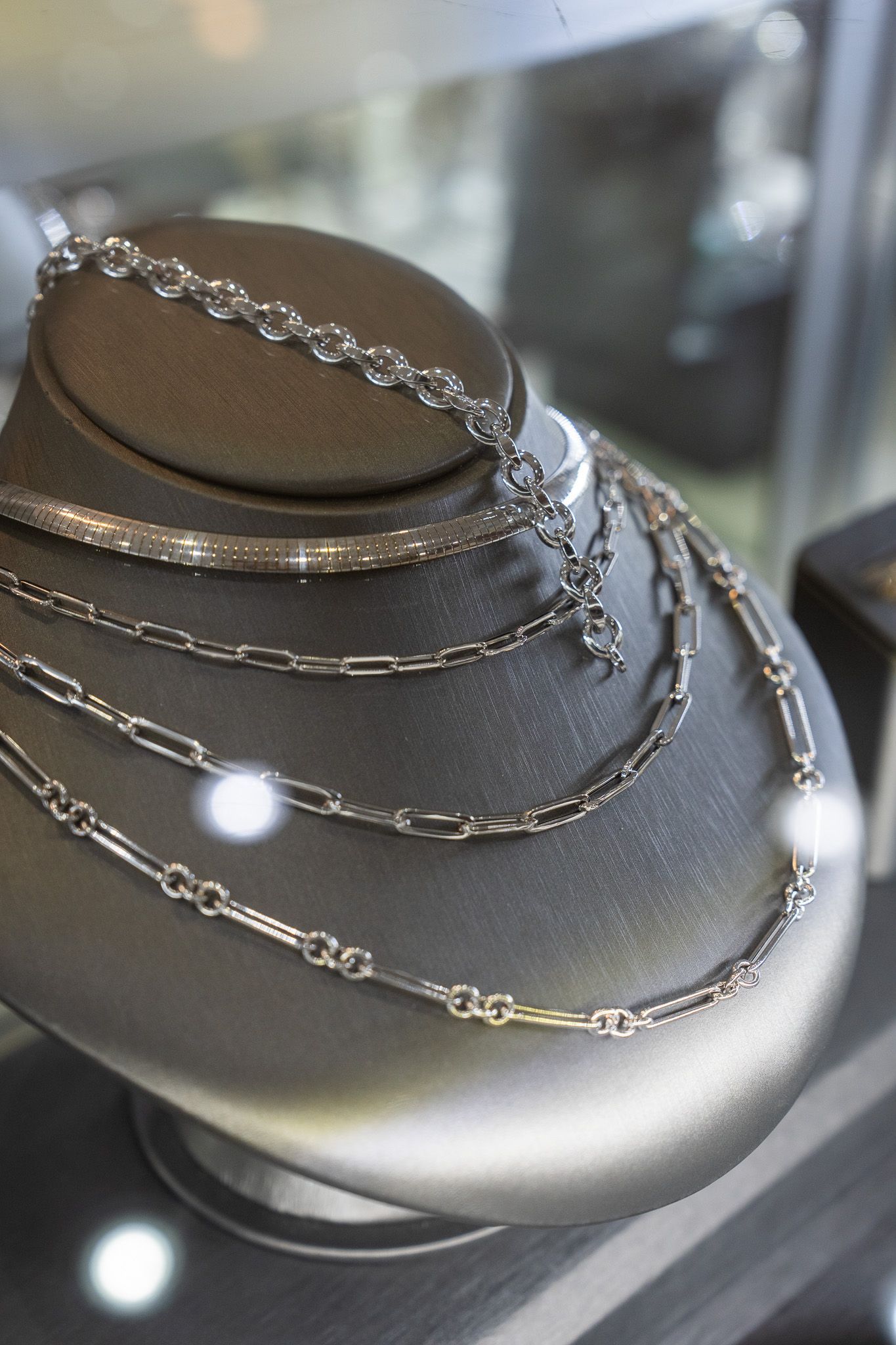 Silver chain necklaces displayed on a black jewelry bust in a glass case.