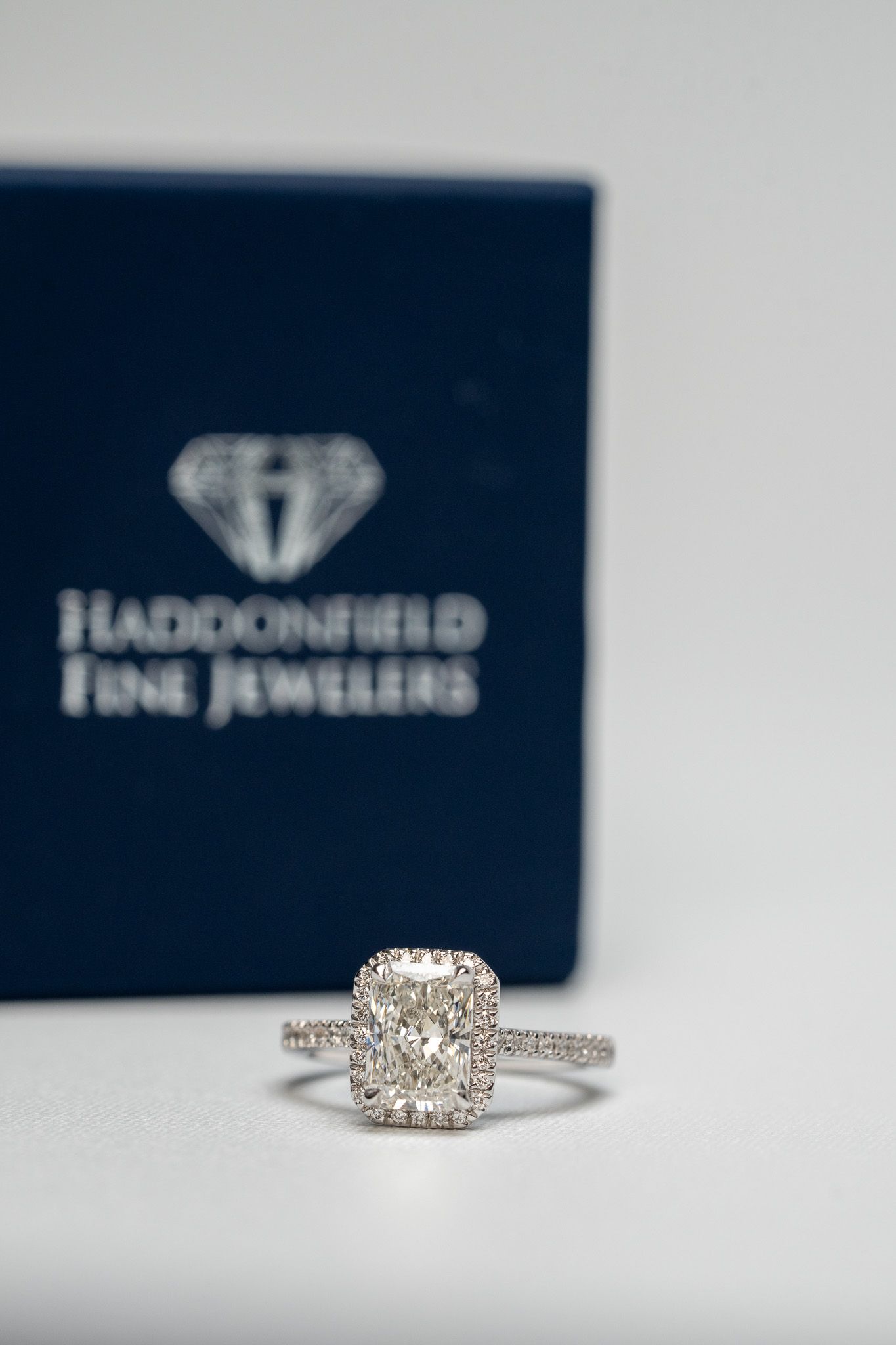 Diamond engagement ring on a white surface, with a blurred blue Haddonfield Fine Jewelers box behind it.