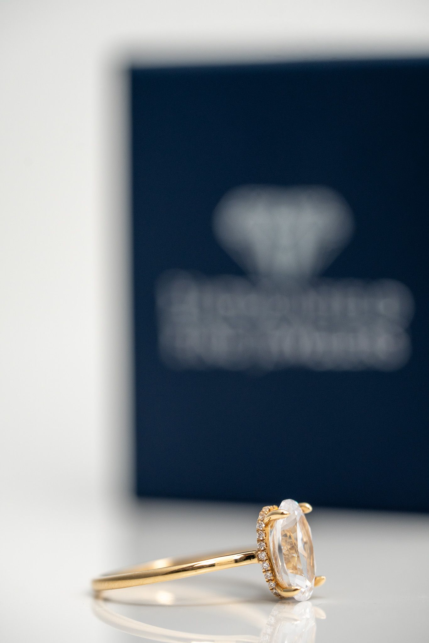 Gold ring with a pear-shaped diamond on a white surface, blurred jewelry box in the background