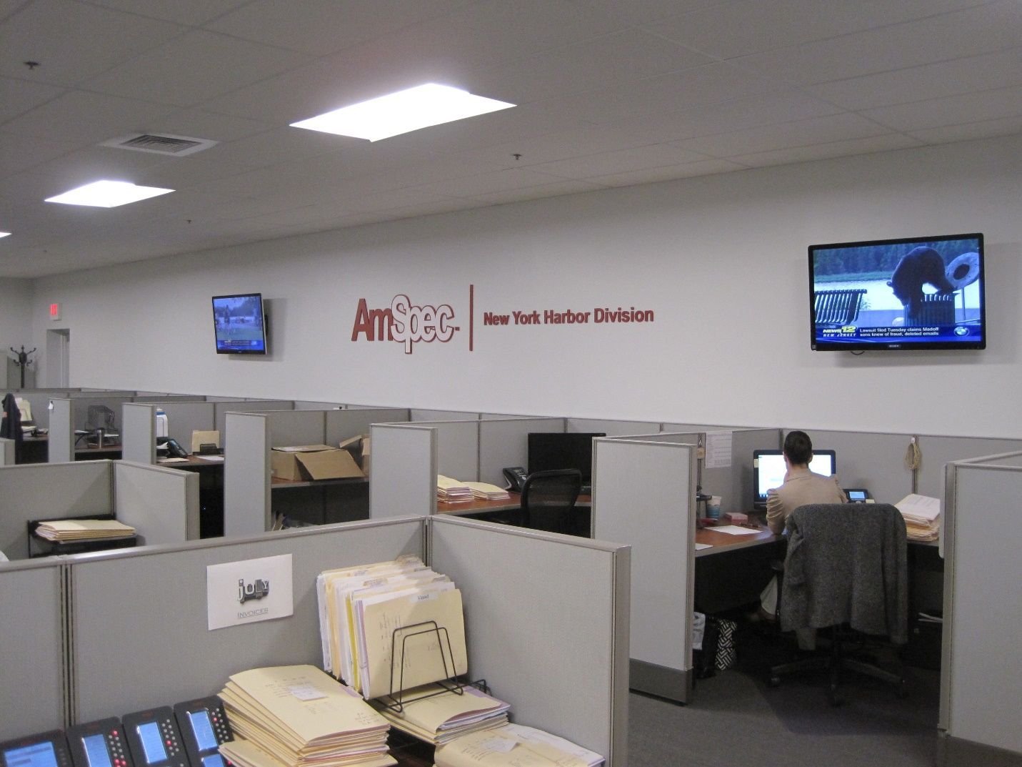 A man sits at a desk in a cubicle in an office with a sign that says amsec.