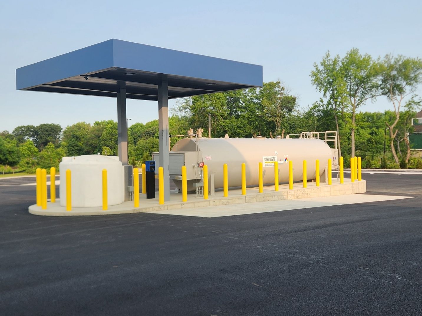 Fuel storage tank under a blue canopy with yellow bollards in an asphalt lot, with a treeline in the background.
